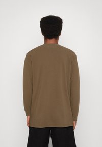 Ruskea pitkähihainen paita, jossa on pyöreä pääntie, sileä pinta ja suora leikkaus. Paidassa on resorit hihansuissa ja pieni suorakaiteen muotoinen lappu takana.