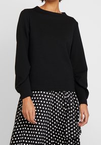 Svart långärmad topp tillverkad av mjukt material med en rundad halsringning. Bärs över en svart och vit prickig kjol med volanger.