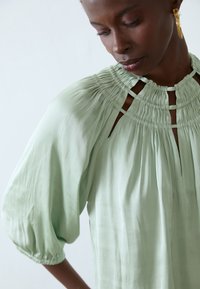 Bluse med puffede ærmer i lys grøn farve har samlet smock ved halsudskæringen og udskæringsdetaljer. Blødt, glat stof med en flydende design.