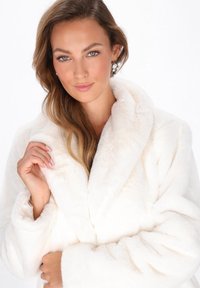 Donna con lunghi capelli ondulati che indossa orecchini a grappolo di perle, tiene il colletto di un soprabito in pelliccia sintetica bianca morbida contro uno sfondo bianco.