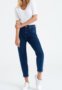 Modré džíny s vysokým pasem a zúženými nohavicemi, v kombinaci s těsným bílým tričkem a bílými teniskami, na jednobarevném pozadí.