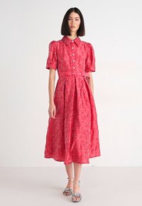 Vestido rojo, hasta la rodilla, con mangas abullonadas, cuello y cinturón en la cintura. La tela presenta bordados florales y una textura suave. Sandalias plateadas.