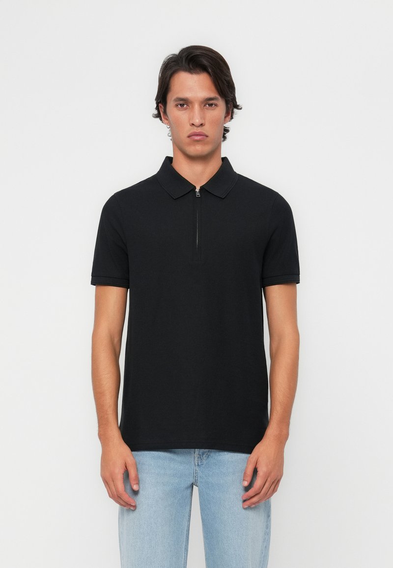 Melna īsām piedurknēm polo krekls ar rāvējslēdzēja apkakli, izgatavots no gluda auduma. Piegriezums ir pieguļošs, minimālistisks dizains, bez redzamām rakstainēm vai akcentiem.