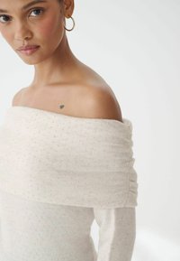 Top off-the-shoulder di colore crema realizzato in morbido tessuto con dettagli di scintillio sottili, caratterizzato da maniche arricciate e una superficie liscia.