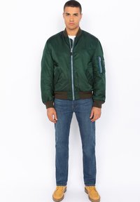 Giacca bomber verde in satin con polsini e fondo a coste, due tasche frontali, una tasca con zip sull'avambraccio e un dettaglio della zip blu. Indossata con jeans blu.