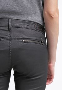 Černé úzké džíny s hladkým povrchem, zipovou boční kapsou a hnědou koženou nášivkou se značkou na zadním pásku.