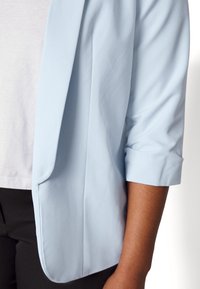 Light blue blazer with three-quarter sleeves worn over a white shirt and black pants, shown from chest to mid-thigh.