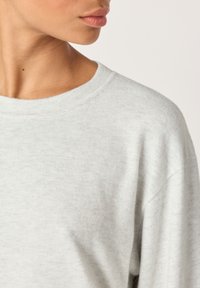 Närbild av en persons nacke och nedre ansikte som bär en ljusgrå tröja med rund hals mot en neutral bakgrund.