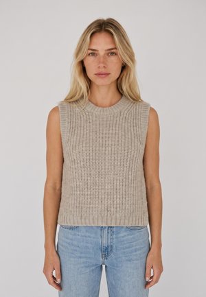 Mujer rubia con un jersey de punto sin mangas beige y vaqueros azul claro sobre un fondo liso y claro.