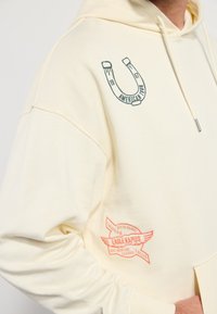 Cremefarvet hoodie med en afslappet pasform; har trykte detaljer af en hestesko og tekst på venstre bryst og højre ærme. Blødt stof.