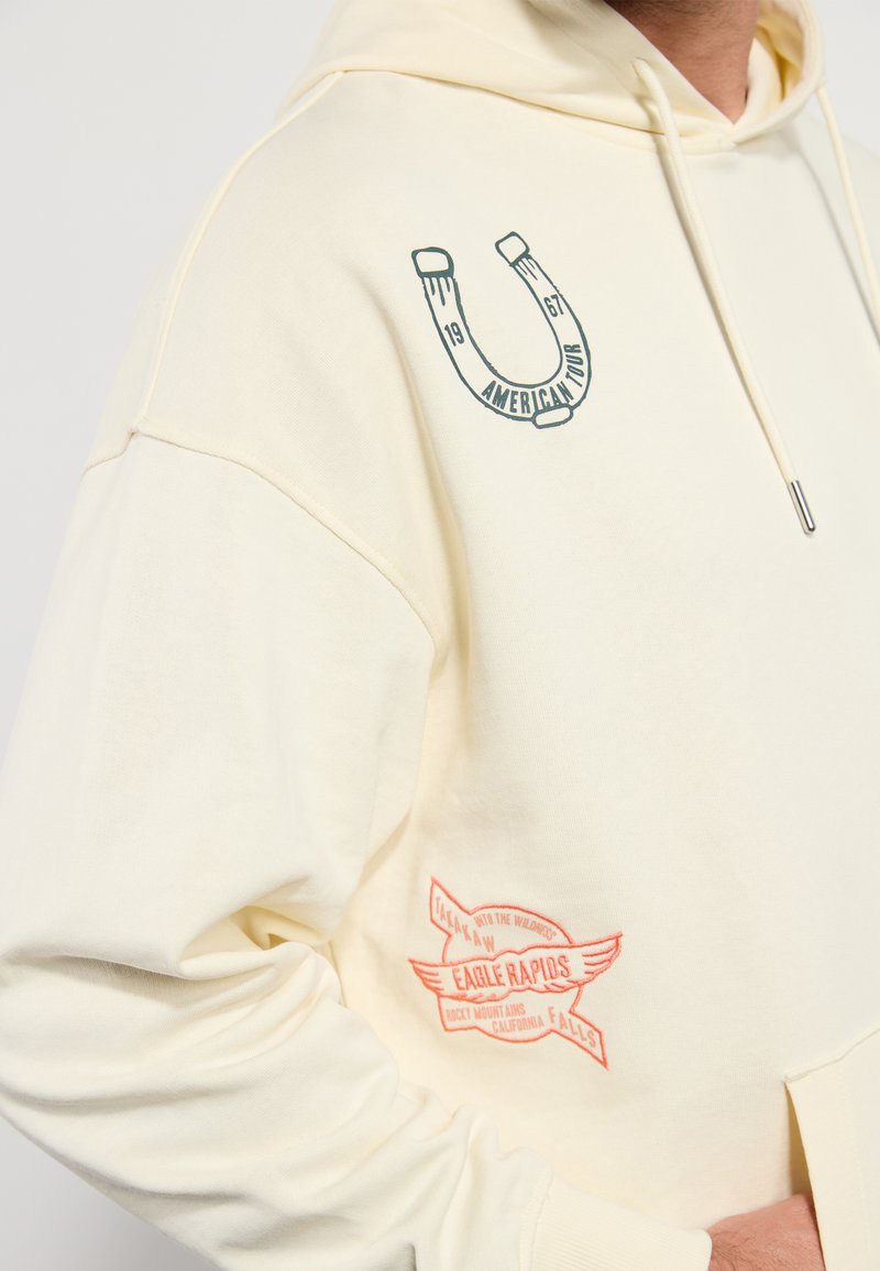 Sweat à capuche couleur crème avec une coupe décontractée ; présente des accents imprimés d'un fer à cheval et de texte sur la poitrine gauche et la manche droite. Tissu doux.