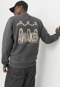 Mørkegrå sweatshirt med et stort grafisk print på ryggen med teksten "AWAKEN" og tre stiliserede figurer. Ribbet manchetter og kant.