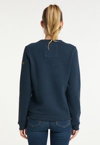 Sweatshirt bleu marine en coton, avec un col rond, des poignets côtelés et un petit patch dans le dos. Porté avec un jean ajusté.
