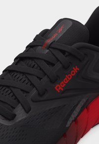 Μαύρο και κόκκινο αθλητικό παπούτσι Reebok με μαύρα κορδόνια και κόκκινο λογότυπο στη πλευρά και στη γλώσσα, με υφή από πλεκτό δίχτυ και τμηματοποιημένη κόκκινη σόλα.