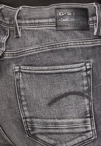 G-Star RAW farkut haalistuneessa harmaassa, nahkatekstillä. Ominaisuuksiin kuuluu takatasku ja erottuva tikkaus taskun reunoilla.