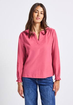 Donna con lunghi capelli castani che indossa una blusa rosa a maniche lunghe con polsini arricciati e jeans blu, in piedi davanti a uno sfondo bianco uniforme.
