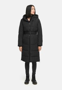 Doudoune noire avec capuche et taille ceinturée. Présente des sections matelassées, une fermeture éclair et des poches latérales. Portée avec des leggings noirs et des bottes.
