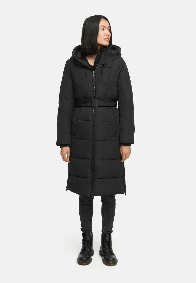 Abrigo de plumas negro con capucha y cintura ceñida. Presenta secciones acolchadas, cierre con cremallera y bolsillos laterales. Llevado con legging negros y botas.