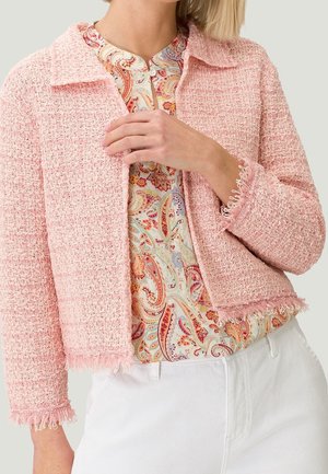 Femme portant une veste courte texturée rose par-dessus un chemisier à motif cachemire et un pantalon blanc, ajustant le devant de la veste avec la main.