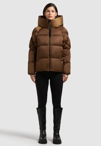 Braune Steppjacke mit hohem, gepolstertem Kragen, horizontaler Steppung und Reißverschluss. Getragen über schwarzen Leggings und hohen schwarzen Stiefeln.