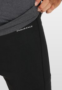 Zwarte sportbroek met een zachte textuur, voorzien van een aansluitend ontwerp en zijnaaddetails. Het merk "ENDURANCE" is in het wit op de tailleband gedrukt.