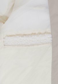 Veste légère en tissu blanc à texture lisse avec un accent beige sur l'épaule, incluant une poche doublée en fourrure.