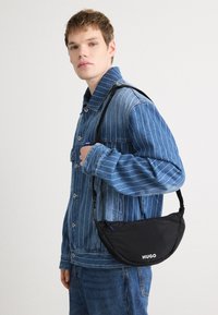 Bolsa preta com uma forma semicircular e fecho de zíper, ostentando o logótipo da marca "HUGO" em branco. Usada sobre uma jaqueta de ganga.