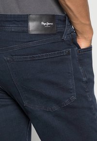 Mörkblå denimjeans med vävd textur, logotyppatch på midjebandet och framträdande sömmar på bakfickorna med ett V-mönster.