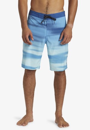 Modèle masculin portant un short de bain dégradé bleu avec un cordon noir, debout pieds nus devant un fond blanc uni.
