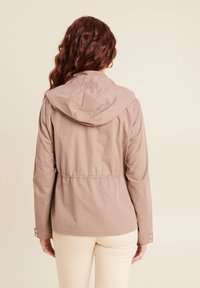 Giacca marrone chiaro con cappuccio, vita elastica e polsini con bottoni. Texture liscia con dettagli di cucitura minimi. Indossata sopra pantaloni color crema.