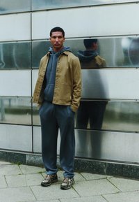 Tenue décontractée composée d'une veste marron à boutons portée sur un sweat à capuche gris, associée à un pantalon de jogging gris et des baskets marron, le tout sur un fond métallique.