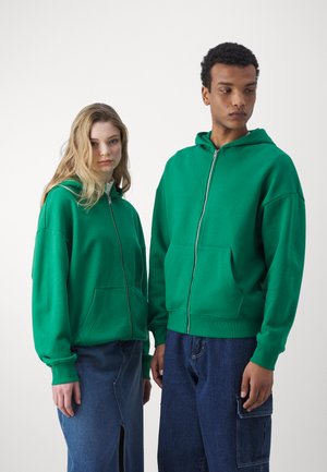 Grønne zip-up hættetrøjer med frontlommer, lavet af blødt stof. Begge modeller bærer afslappede denim-bund, med en løs pasform og synlig syning.