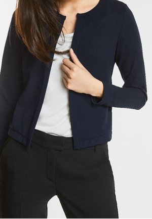 Frau trägt eine marineblaue, offen vorne getragene Strickjacke über einem weißen Oberteil und schwarzen Hosen, Hand hält die Strickjacke nahe an der Brust.