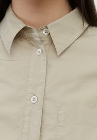 Camicia beige con colletto e bottoni bianchi, dotata di taschino sul petto. Il tessuto appare liscio con texture minima.