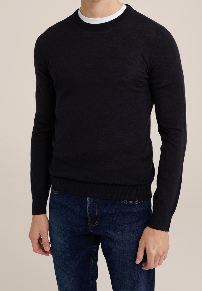 Homme portant un pull à manches longues noir par-dessus un sous-vêtement blanc et un jean bleu foncé, debout devant un fond neutre.