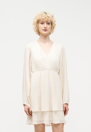 Žena s krátkymi kučeravými blond vlasmi, ktorá má na sebe biele plisované šaty s dlhým rukávom a výstrihom do V na jednoduchom pozadí.
