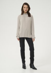 Light beige high-neck blouse with elastic gathering, long sleeves, and a smooth texture, paired with black fitted pants and ankle boots.