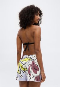 Mujer con cabello rizado, vistiendo un top de bikini de tirantes negro y shorts con un patrón abstracto colorido, de pie de espaldas a la cámara.