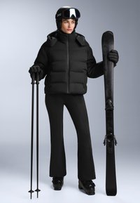 Fekete puffertető, magas gallérral, fekete sínadrág, fekete sícipők, fekete síbotok és kötések, fekete sisakkal és szemüveggel párosítva.