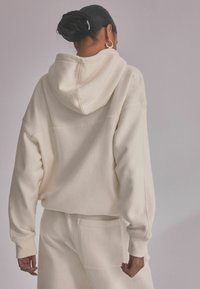 Light beige hoodie with a large hood, dropped shoulders, and ribbed cuffs. Material appears soft and textured, styled in a relaxed fit.