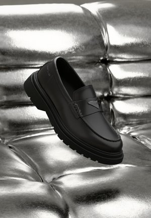 Svarte skinn slip-on sko med avrundet tå, strukturert søm og gummisåle. Plassert på en blank, sølvfarget tuftet overflate.