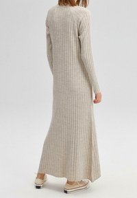 Robe tricotée beige à côtes, longue, avec manches longues et encolure arrondie, présentant un ourlet légèrement évasé et des fentes sur les côtés, accompagnée de chaussures claires.