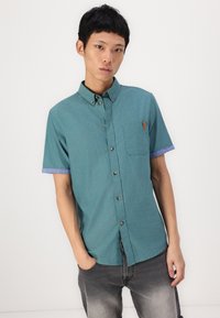 Chemise à manches courtes avec boutons en tissu teal, dotée d'une poche poitrine et de poignets bleu. Texturée, avec des boutons en métal marron.