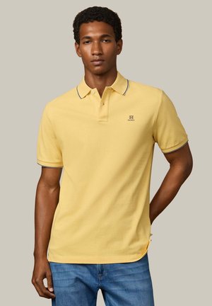 Joven vestido con una camiseta polo amarilla de manga corta con borde negro y pantalones vaqueros azules, de pie contra un fondo neutro y liso.