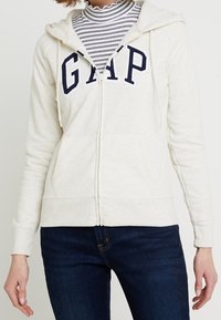 Sudadera con cremallera color crema con el logo "GAP" en azul marino, capucha con cordón, bolsillos frontales y puños acanalados. Llevada sobre un cuello de tortuga a rayas.