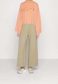 Cropped, textured orange sweater with a swirl logo, paired with wide-legged beige pants and light brown sneakers.
