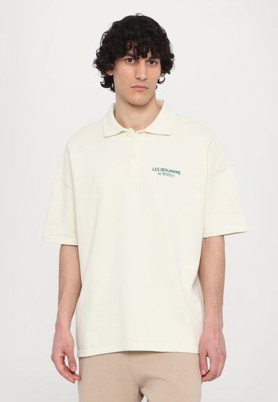 Polo shirt beige surdimensionné avec un col, portant un logo brodé vert sur la poitrine gauche. Tissu doux, manches courtes et coupe décontractée.