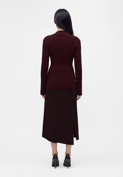 Femme aux longs cheveux noirs portant un pull côtelé foncé et une jupe sombre assortie avec une fente latérale, associée à des chaussures à talons noires, debout de dos.