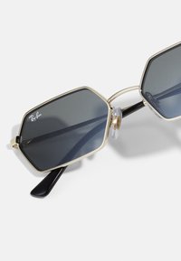 Kuusikulmion muotoiset aurinkolasit tummilla linsseillä, kultaisissa metallikehyksissä ja mustissa asetaattioksilla. Varustettu kirkkailla nenätyynyillä ja Ray-Ban -logolla linssissä.