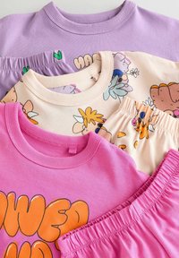 Vier gestapelde kledingstukken: lavendelkleurige crewneck, licht perzikkleurig met bloemenprint, roze met oranje tekst, en elastische roze shorts. Zacht katoenen stof.